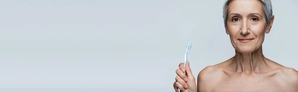 Весела жінка середнього віку, що тримає зубну щітку ізольовано на сірому, банер — стокове фото