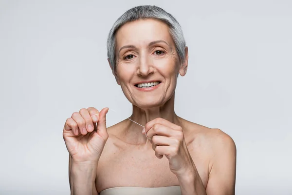 Mujer feliz de mediana edad con pelo gris sosteniendo hilo dental aislado en gris - foto de stock