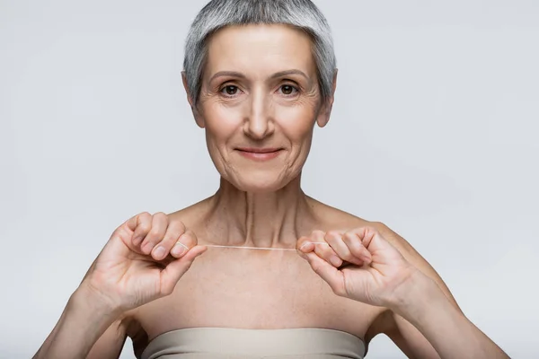 Весёлая женщина средних лет с седыми волосами, держащая зубную нить, изолированную на серой — стоковое фото