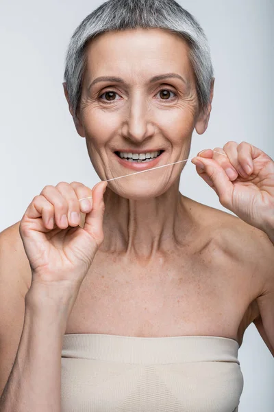 Довольная женщина средних лет с седыми волосами, держащая зубную нить и улыбаясь изолированно на серой — стоковое фото