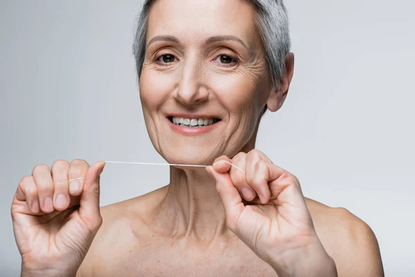 Mujer de mediana edad complacida con pelo gris sosteniendo hilo dental y sonriendo aislado en gris - foto de stock