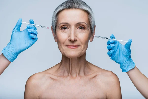 Beauticians in latex gloves holding syringe with beauty injection near smiling middle aged client isolated on grey — Stock Photo