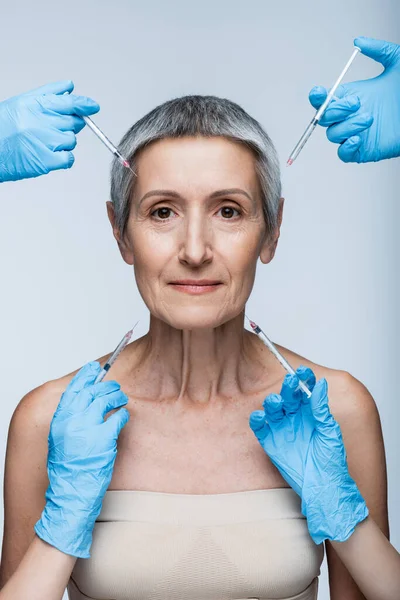 Beauticians in blue latex gloves holding syringe with beauty injection near middle aged client isolated on grey — Stock Photo