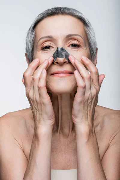 Весела зріла жінка наносить чорну пляму на ніс ізольовано на сірому — стокове фото
