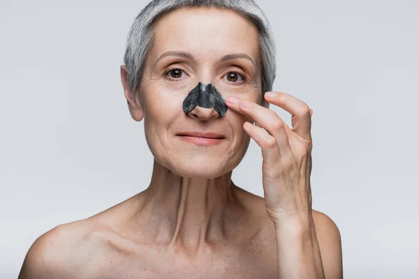 Femme positive et mature appliquant tache noire sur le nez isolé sur gris — Photo de stock