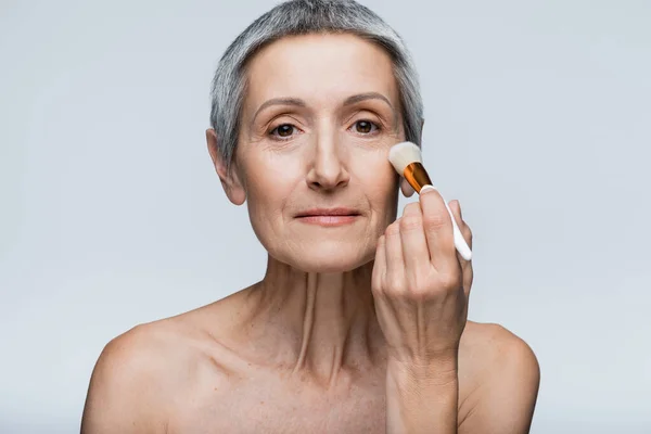 Mature woman applying face powder with cosmetic brush and looking at camera isolated on grey — Stock Photo