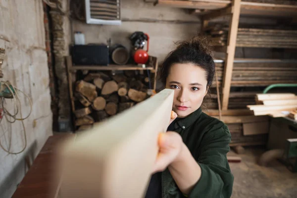 Brunette carpenter checking wooden plank in blurred workshop — Stock Photo