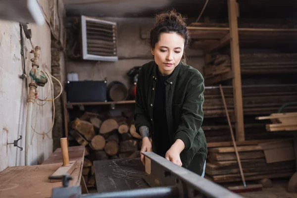 Татуированный плотник, работающий на соединительной машине возле размытых деревянных досок в мастерской — стоковое фото
