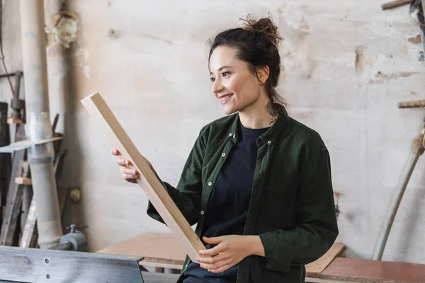 Trabajador de madera positivo sosteniendo tablón cerca de la máquina de unión en el taller - foto de stock