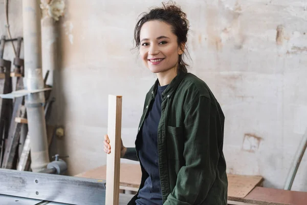 Junger Tischler hält Holzplanke in der Werkstatt und lächelt in die Kamera — Stockfoto