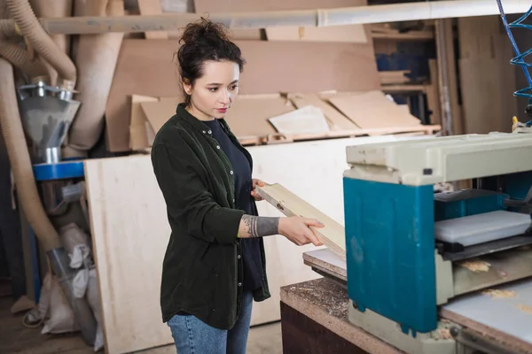 Tattooed woodworker holding plank near thickness planer in workshop — Stock Photo