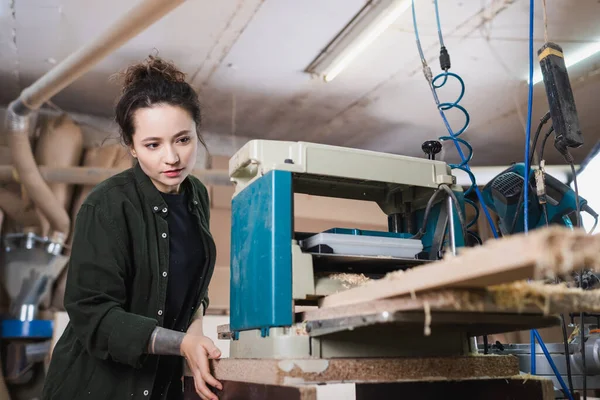 Brunette woodworker looking at blurred plank in thickness planer — Stock Photo