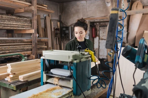 Jeune menuisier debout près du banc épaississeur et planches en bois dans l'atelier — Photo de stock