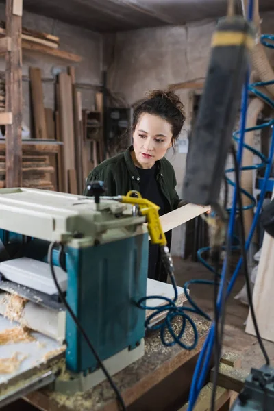 Brunette carpenter holding wooden board near thickness planer in workshop — Stock Photo