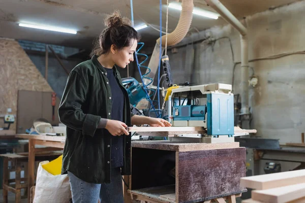 Vista laterale del falegname che tiene il bordo di legno vicino alla pialla di spessore in officina — Foto stock