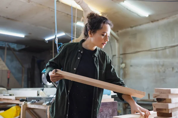 Jeune charpentier tenant le conseil près des planches en bois dans l'atelier — Photo de stock