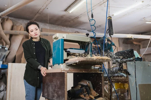 Giovane falegname guardando segatura sfocata vicino spessore pialla in officina — Foto stock