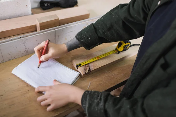 Vue recadrée de l'écriture du menuisier sur carnet près de la planche de bois et ruban à mesurer dans l'atelier — Photo de stock