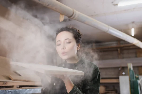 Charpentier soufflant la sciure de bois de la planche en bois dans l'atelier — Photo de stock