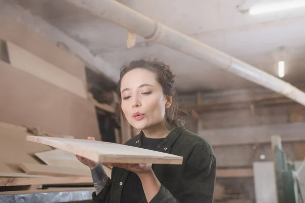 Brünetten Tischler pustet Staub von Holzplanke in Werkstatt — Stockfoto