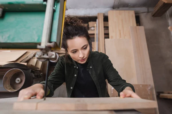 Diseñador de muebles morena sosteniendo borrosa cerca de la sierra de banda en el taller - foto de stock