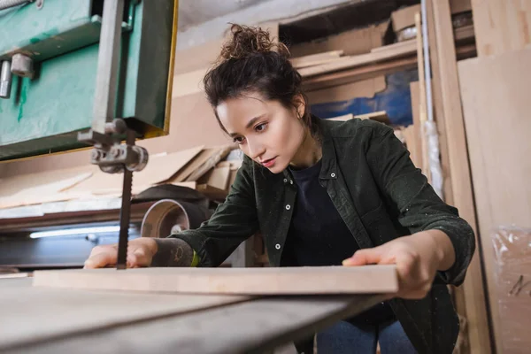 Giovane falegname con tavola di legno vicino alla sega a nastro sfocata in officina — Foto stock