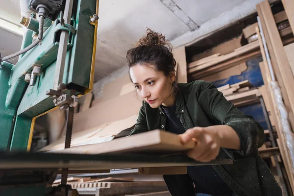 Brunette charpentier tenant une planche floue près de la scie à ruban en atelier — Photo de stock