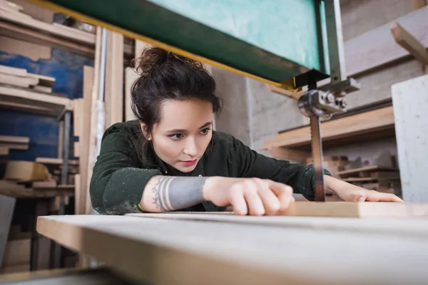 Brunette menuisier tenant une planche de bois floue et travaillant dans une scie à ruban en atelier — Photo de stock