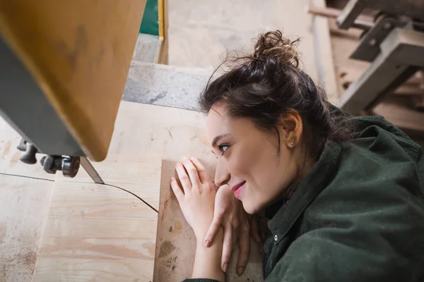 Vue latérale du concepteur de meubles souriants regardant la planche et la scie à ruban dans l'atelier — Photo de stock