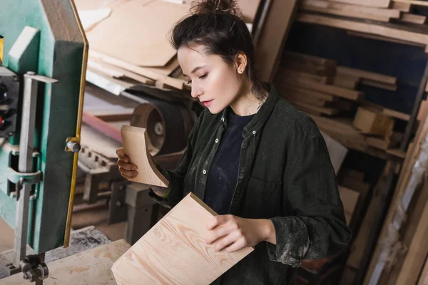 Молодой плотник в опилках держит доски возле ленточной пилы в мастерской — стоковое фото