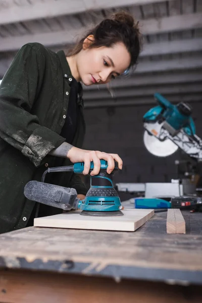 Charpentier flou à l'aide d'une ponceuse lors du polissage de la planche en atelier — Photo de stock