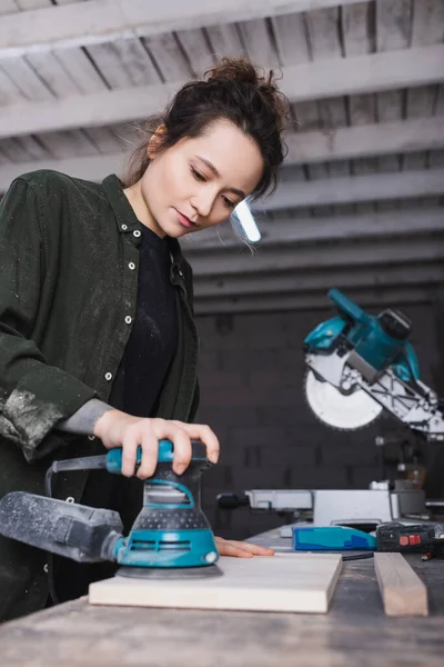 Carpenter using blurred sander on blurred plank near miter saw in workshop — Stock Photo