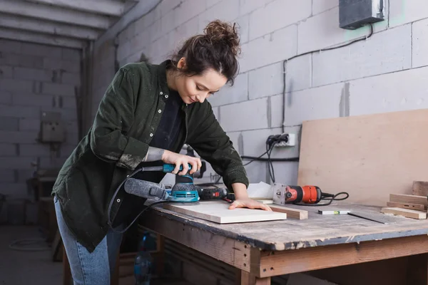 Carpintero en camisa lijado tablón de madera en taller - foto de stock