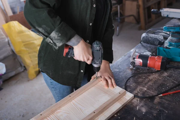 Vue recadrée du concepteur de meubles travaillant avec perceuse électrique et planche en bois dans l'atelier — Photo de stock