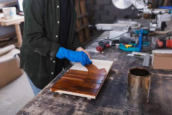 Vista recortada del diseñador de muebles en guante de goma aplicando una mancha de madera a bordo en el taller - foto de stock