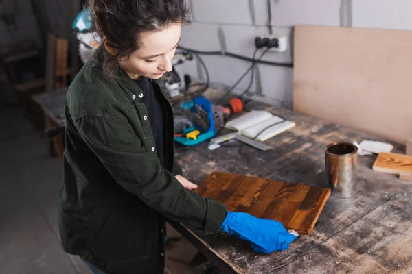 Brünette Handwerkerin trägt in verschwommener Werkstatt Holzbeize auf Bord auf — Stockfoto