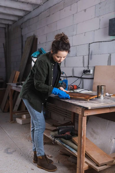 Young carpenter in rubber glove applying wood stain on plank near jar and miter saw in workshop — Stock Photo