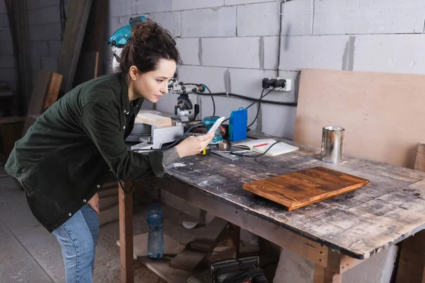 Falegname scattare foto su smartphone vicino a bordo di legno in officina — Foto stock