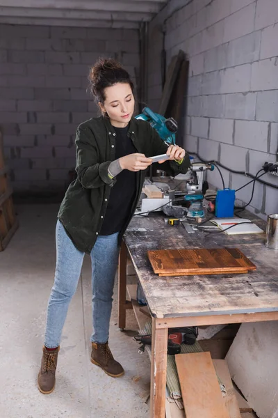 Carpintero tatuado tomando fotos en smartphone cerca de tablón de madera en taller - foto de stock