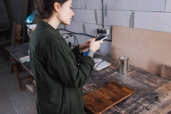 Seitenansicht einer jungen Handwerkerin mit Smartphone in der Nähe von Holzplanke in Werkstatt — Stockfoto