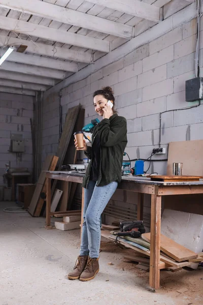 Lächelnder Möbeldesigner hält Coffee to go in der Werkstatt und spricht mit Smartphone — Stockfoto