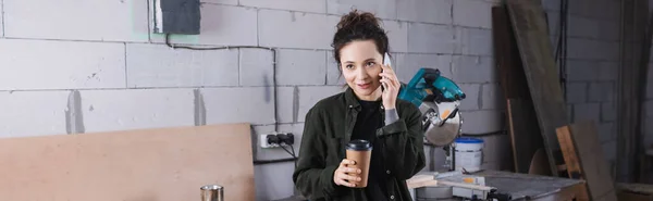 Positive carpenter holding paper cup and talking on smartphone in workshop, banner — Stock Photo