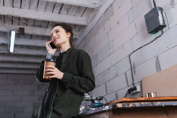 Low angle view of cheerful carpenter talking on mobile phone and holding coffee to go in workshop — Stock Photo