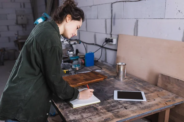 Vue latérale de l'écriture du menuisier sur ordinateur portable près de la tablette numérique avec écran blanc dans l'atelier — Photo de stock