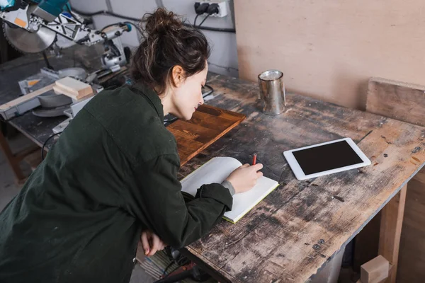 Hohe Winkelansicht des Tischlers Schreiben auf Notizbuch in der Nähe von digitalem Tablet und Holzbeize in der Werkstatt — Stockfoto