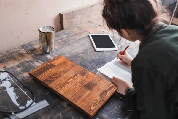 Concepteur de meubles flous écrivant sur un carnet près d'une planche de bois et d'une tablette numérique — Photo de stock