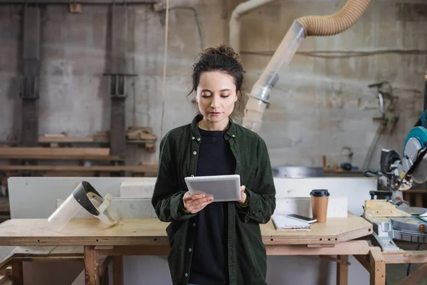 Jeune menuisier utilisant une tablette numérique près de la visière de sécurité et du café pour aller en atelier — Photo de stock