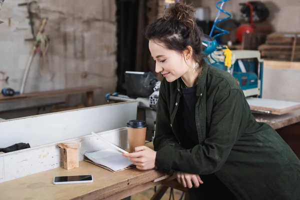 Junger Schreiner mit digitalem Tablet in der Nähe von Notebook und Coffee to go in Werkstatt — Stockfoto