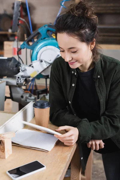 Trabajador de la madera sonriente usando tableta digital cerca del teléfono celular y la taza de papel en el taller - foto de stock