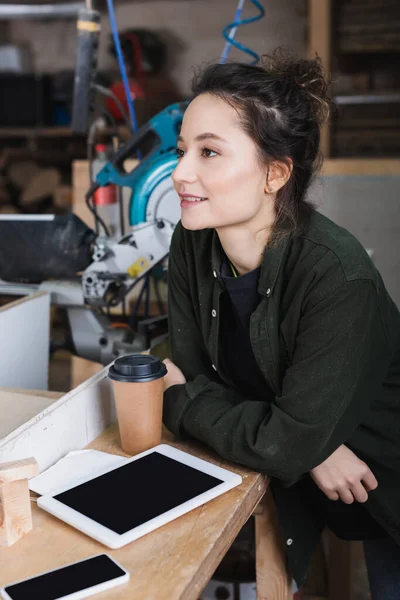 Glücklicher Tischler steht neben Geräten mit leerem Bildschirm und Kaffee to go im Büro — Stockfoto